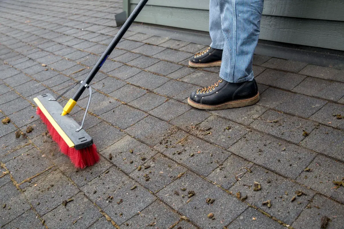 A push broom sweeping loosened moss off asphalt shingles beside the house siding